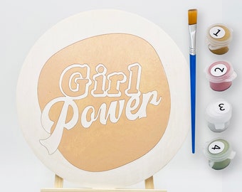 Könnte beinhalten: Eine runde weiße Holzleinwand mit goldenem Hintergrund und dem Text "Girl Power" in Weiß. Die Leinwand steht auf einem Holzstaffelei. Es gibt vier kleine Farbdosen mit den Nummern 1 bis 4.