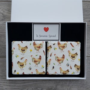 May include: A set of two square coasters featuring a white background with a watercolour pattern of brown chickens, yellow flowers, and white feathers. The coasters are presented in a white box with a black interior and a small card that reads "To Someone Special" with a red heart.