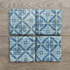 May include: Set of four blue and white ceramic coasters with a geometric pattern. The coasters have a white background with blue and gold accents.