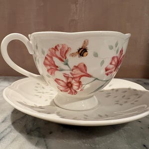 May include: White teacup and saucer set with a floral design featuring pink flowers and a bumblebee. The teacup has a curved handle and sits on a matching saucer with a scalloped edge. A delicate and charming tea set.