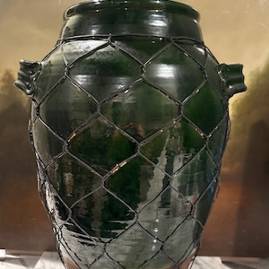 May include: A large, dark green glazed ceramic vase with a wire mesh overlay. The vase has a wide body, a narrow neck, and two small handles. The wire mesh is dark and adds a decorative element to the vase.