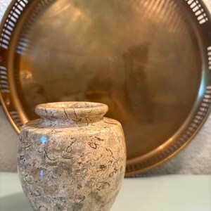 Puede incluir: Un peque&ntilde;o jarr&oacute;n decorativo con un patr&oacute;n moteado beige y verde. El jarr&oacute;n est&aacute; frente a una gran bandeja redonda de color dorado con un borde decorativo. La bandeja est&aacute; ligeramente desenfocada.