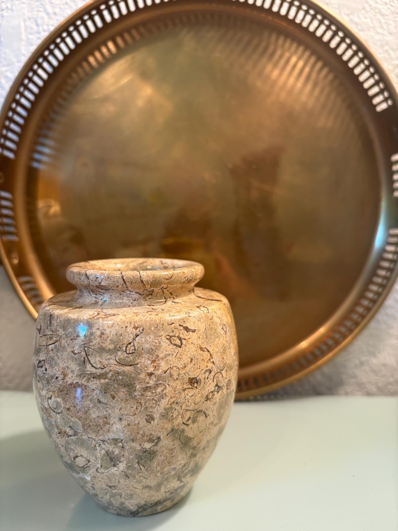 Puede incluir: Un peque&ntilde;o jarr&oacute;n decorativo con un patr&oacute;n moteado beige y verde. El jarr&oacute;n est&aacute; frente a una gran bandeja redonda de color dorado con un borde decorativo. La bandeja est&aacute; ligeramente desenfocada.
