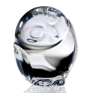 Puede incluir: Una figura de cristal transparente con forma de b&uacute;ho y cuerpo liso y redondeado. El dise&ntilde;o presenta dos hendiduras circulares para los ojos y una sutil forma de pico, con la luz reflej&aacute;ndose en la superficie pulida y transparente.