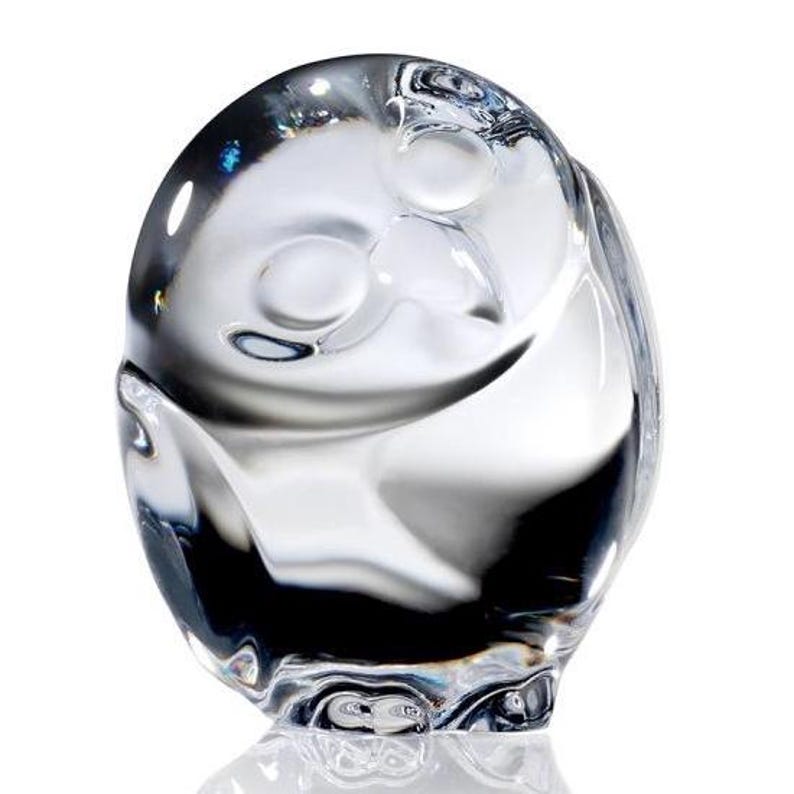 Puede incluir: Una figura de cristal transparente con forma de b&uacute;ho y cuerpo liso y redondeado. El dise&ntilde;o presenta dos hendiduras circulares para los ojos y una sutil forma de pico, con la luz reflej&aacute;ndose en la superficie pulida y transparente.