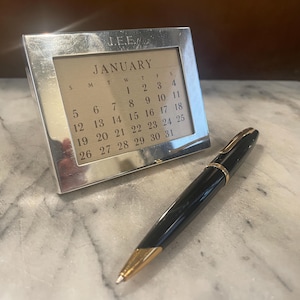 May include: A silver desk calendar with a gold-colored face showing the month of January. The calendar is sitting on a white marble surface next to a black and gold pen.