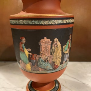 May include: A terracotta vase with a black background and a painted scene of people around a fountain. The vase has a gold trim and a base with a blue and gold design.