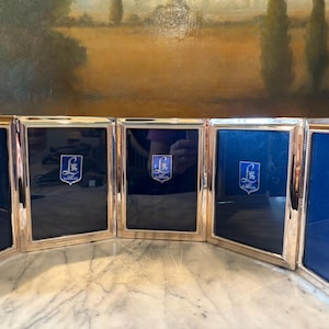May include: A set of five silver-toned picture frames with a blue interior. Each frame features a decorative emblem in the center. The frames are arranged in a row, with a marble surface in the foreground and a painting in the background.