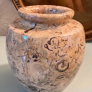 Puede incluir: Un peque&ntilde;o jarr&oacute;n decorativo con un cuerpo redondeado y una abertura estrecha. El jarr&oacute;n est&aacute; hecho de un material similar a la piedra con un patr&oacute;n moteado de beige, marr&oacute;n y gris. La superficie tiene un acabado pulido y el dise&ntilde;o general es simple y elegante.