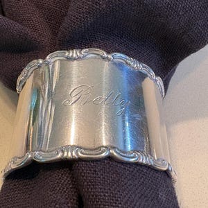 May include: A silver-toned metal napkin ring with a polished finish and decorative scalloped edges. The center of the band features the engraved name Betty in a cursive script. A dark charcoal-colored fabric napkin is threaded through the ring.