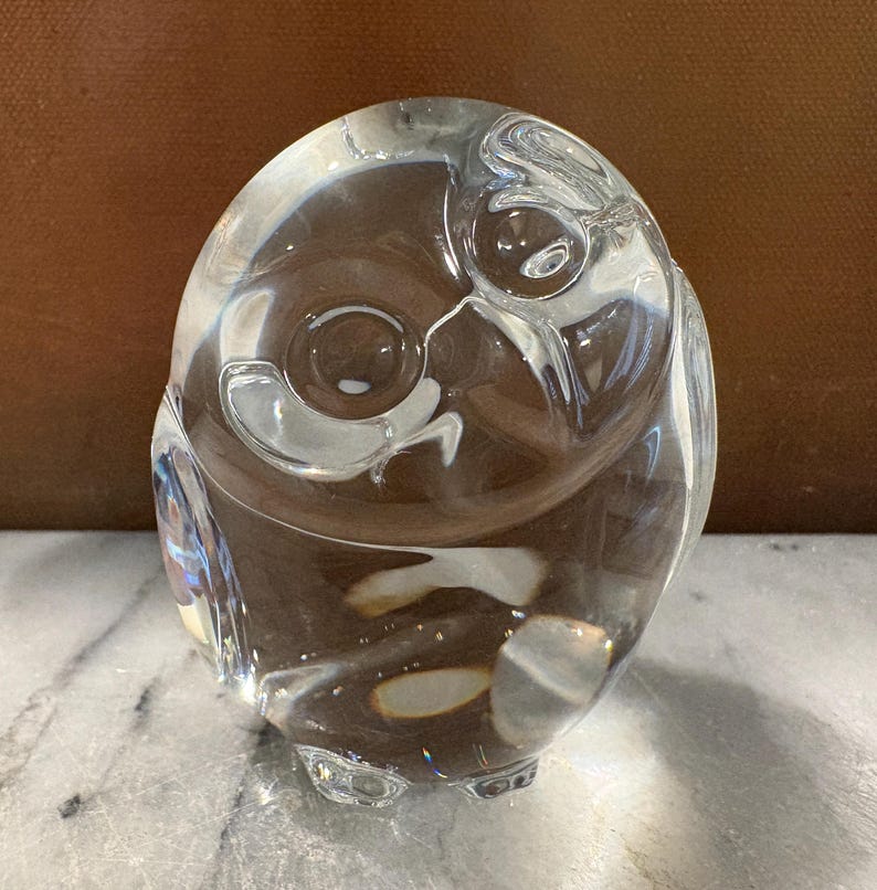Puede incluir: Una figura de cristal transparente con forma de b&uacute;ho y cuerpo liso y redondeado. El dise&ntilde;o presenta dos hendiduras circulares para los ojos y una forma de pico sutil, con una superficie pulida y reflectante que capta la luz y crea reflejos.