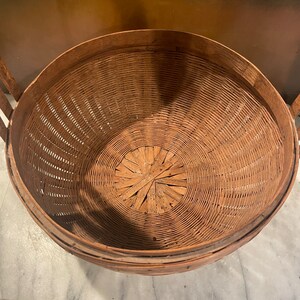 Puede incluir: Una cesta redonda de mimbre marr&oacute;n tejida con una abertura ancha y un fondo plano. La cesta est&aacute; hecha de materiales naturales y tiene un aspecto r&uacute;stico y vintage.