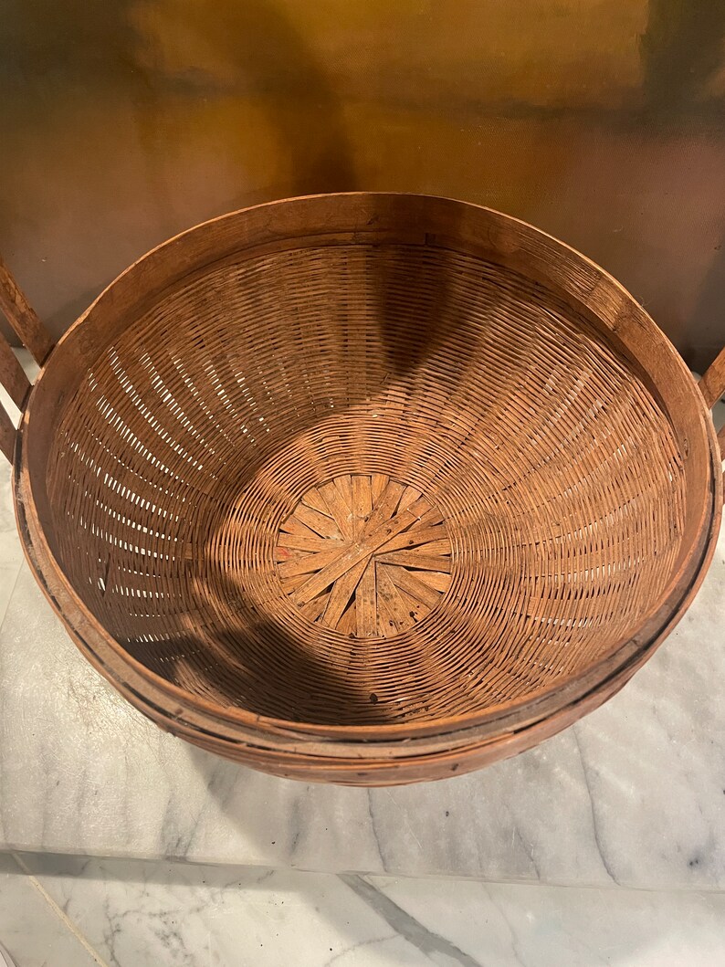 Puede incluir: Una cesta redonda de mimbre marr&oacute;n tejida con una abertura ancha y un fondo plano. La cesta est&aacute; hecha de materiales naturales y tiene un aspecto r&uacute;stico y vintage.