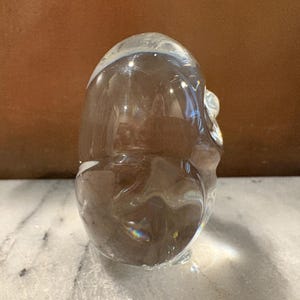 Puede incluir: Una escultura de vidrio transparente y lisa con forma de figura abstracta redondeada. El objeto presenta una superficie pulida y reflectante con sutiles curvas internas y una calidad transparente que permite el paso de la luz, creando reflejos suaves.