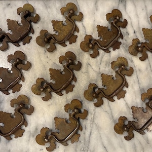 May include: A collection of ten matching brass drawer pulls featuring ornate, scalloped backplates with intricate cutouts and curved, swinging bail handles. Each piece displays a weathered, aged patina with dark brown and muted gold tones.