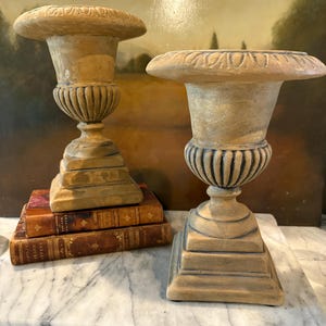 May include: Two decorative urns with a beige finish and a tiered base. The urns are made of a ceramic material and have a classic design. The urns are sitting on a marble surface.
