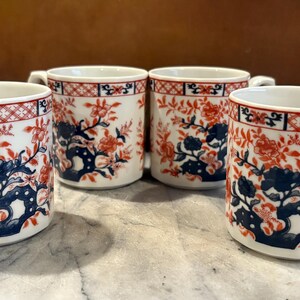 May include: Four white ceramic mugs with ornate handles. Each mug features a repeating pattern of red floral and blue botanical designs. The rims are trimmed with a red and black geometric border.
