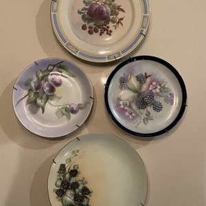May include: Four decorative plates with painted fruit and berry designs. The plates are white with gold trim and feature plums, grapes, blackberries, and flowers.