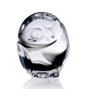 May include: A clear glass figurine shaped like a stylized owl with a rounded body, smooth surface, and subtle indentations representing eyes and a beak. The transparent material reflects light, creating highlights and shadows across the curved form.