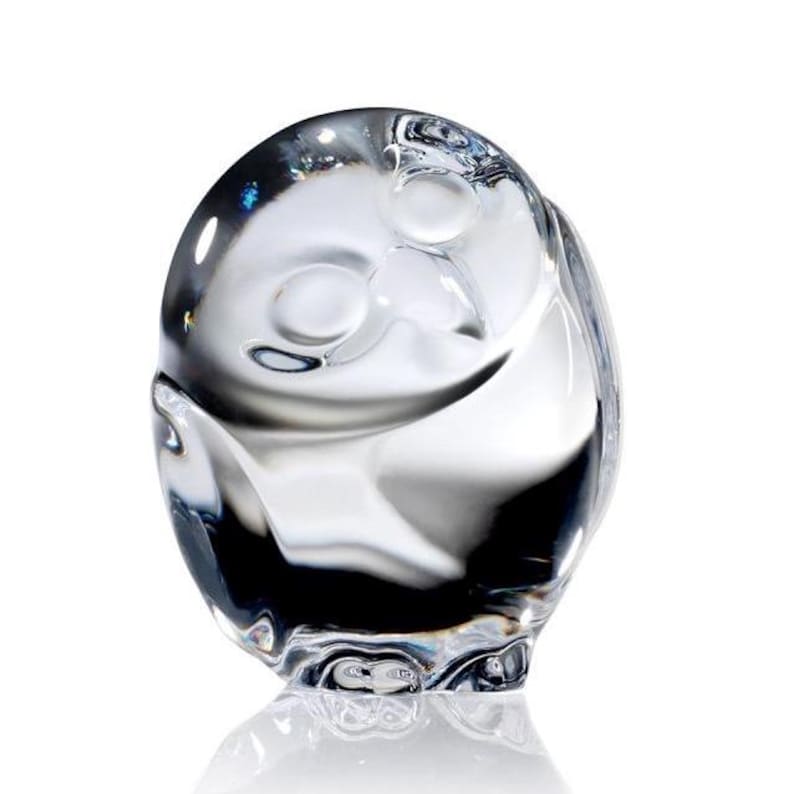Puede incluir: Una figura de cristal transparente con forma de b&uacute;ho estilizado, cuerpo redondeado, superficie lisa y sutiles hendiduras que representan los ojos y el pico. El material transparente refleja la luz, creando brillos y sombras en la forma.