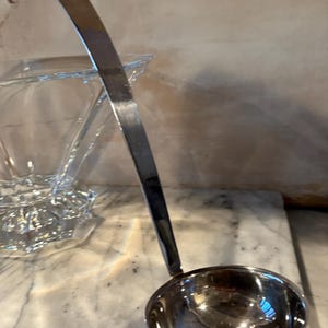 May include: A silver ladle with a long handle and a rounded bowl. The ladle is made of shiny metal and reflects the surrounding light. The kitchen utensil is designed for serving soups and sauces.