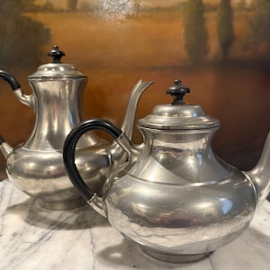 May include: Two antique silver teapots with black handles and black finials. The teapots are displayed on a marble surface. The background features a blurred painting with warm tones.
