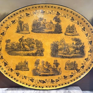 May include: Oval serving tray with a yellow background and black illustrations of people in a garden setting. The tray has a black border with grape vine designs and two handles. A decorative piece for serving.