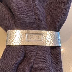 May include: A silver-toned metal napkin ring with a hammered texture and a central rectangular plaque engraved with the letters F.N. The napkin ring is wrapped around a dark purple fabric napkin.