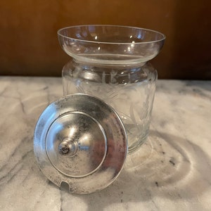 May include: A clear glass jar with an etched floral design and a silver lid. The lid has a small spout for pouring.