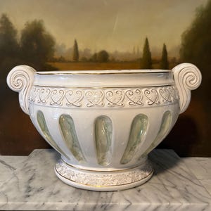 May include: A white ceramic bowl with two scroll-shaped handles, featuring a gold-trimmed rim and a repeating heart-shaped pattern. The body has vertical recessed panels with a pale green iridescent finish, and the base has a gold floral motif.