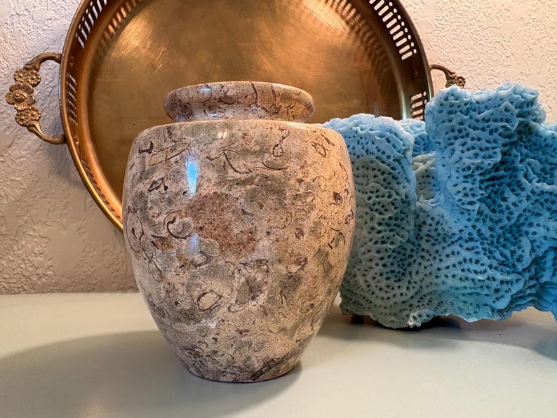 Puede incluir: Un jarr&oacute;n decorativo de piedra moteada con tapa a juego. El jarr&oacute;n es una mezcla de tonos beige, marr&oacute;n y gris. Una bandeja dorada con asas ornamentadas est&aacute; en el fondo, junto con un trozo de coral azul.
