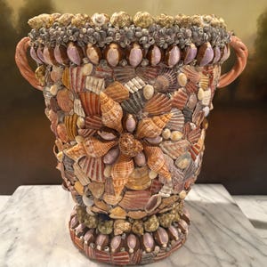 May include: A decorative urn covered in a dense mosaic of various seashells in shades of brown, tan, cream, and purple. The vessel features two curved, peach-colored handles on the sides and a wide rim adorned with small, tightly packed shells.