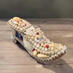 May include: A decorative shoe covered in various seashells, with small red accents. The shoe is beige and tan, with a textured surface. The shoe is displayed on a wooden surface.
