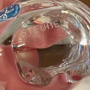 Puede incluir: Un objeto de vidrio transparente con una forma suave y redondeada. La palabra STEUBEN est&aacute; grabada en la superficie del vidrio. Una peque&ntilde;a pegatina azul circular con texto blanco y un icono gr&aacute;fico est&aacute; adherida a la parte superior izquierda.