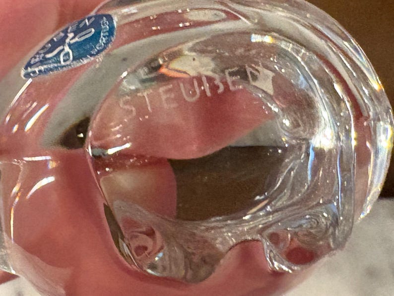 Puede incluir: Un objeto de vidrio transparente con una forma suave y redondeada. La palabra STEUBEN est&aacute; grabada en la superficie del vidrio. Una peque&ntilde;a pegatina azul circular con texto blanco y un icono gr&aacute;fico est&aacute; adherida a la parte superior izquierda.