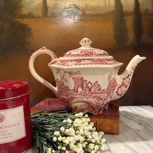 May include: A cream-colored ceramic teapot featuring a red transferware pattern of a castle, bridge, and floral motifs. The teapot has a hexagonal body, a matching lid with a finial, a curved handle, and a spout with floral detailing.