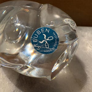 Puede incluir: Un objeto de vidrio transparente con una forma suave, redondeada y org&aacute;nica. Una pegatina azul circular est&aacute; adherida a la superficie, con el texto blanco "STEUBEN" y "MADE IN PORTUGAL" alrededor de un gr&aacute;fico de unas tijeras abiertas.