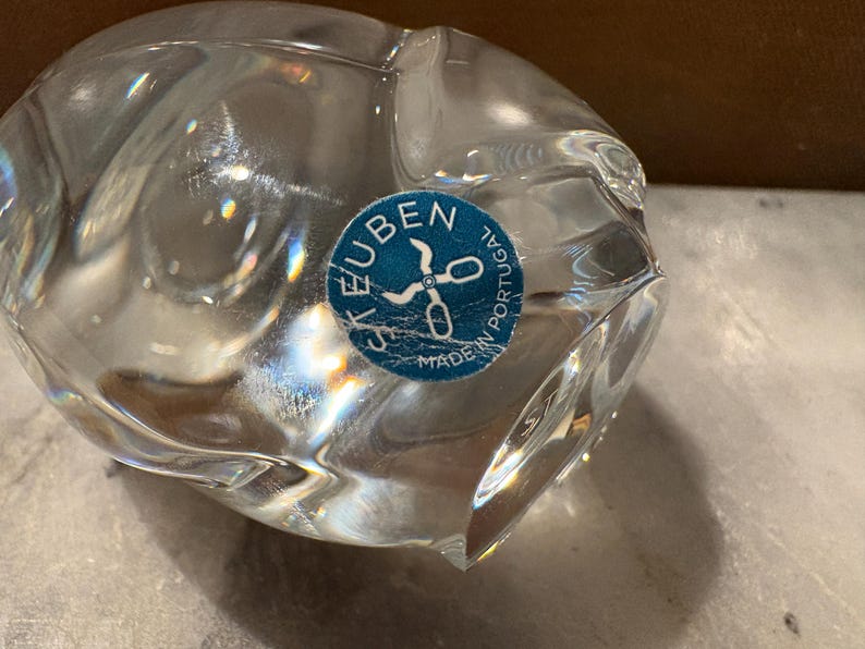 Puede incluir: Un objeto de vidrio transparente con una forma suave, redondeada y org&aacute;nica. Una pegatina azul circular est&aacute; adherida a la superficie, con el texto blanco "STEUBEN" y "MADE IN PORTUGAL" alrededor de un gr&aacute;fico de unas tijeras abiertas.