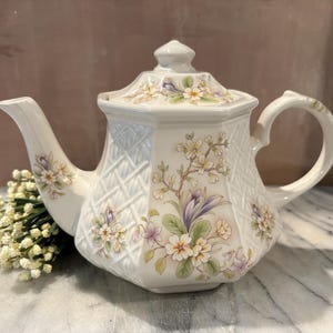 May include: A cream-colored ceramic teapot with an octagonal shape and a lid. The body features a raised lattice texture and a delicate floral pattern with white, yellow, and purple flowers and green leaves. A curved handle and spout are present.