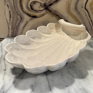 May include: A glossy off-white ceramic dish shaped like a large scallop shell. The dish features a scalloped edge, a curved handle on one side, and a series of embossed radial ridges that extend from the base toward the outer rim of the shell.