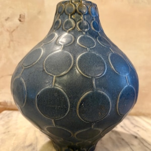 May include: A bulbous ceramic vase with a matte dark blue finish, featuring a repeating embossed pattern of interconnected circles linked by vertical lines. The surface texture appears slightly granular, and the circular motifs are outlined in a lighter, muted tone, creating a structured geometric design across the rounded body and narrow neck.