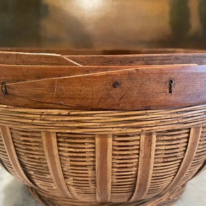 Puede incluir: Una cesta tejida de bamb&uacute; marr&oacute;n y natural con fondo redondeado y una abertura ancha. La cesta tiene un asa a cada lado y est&aacute; hecha de materiales naturales.