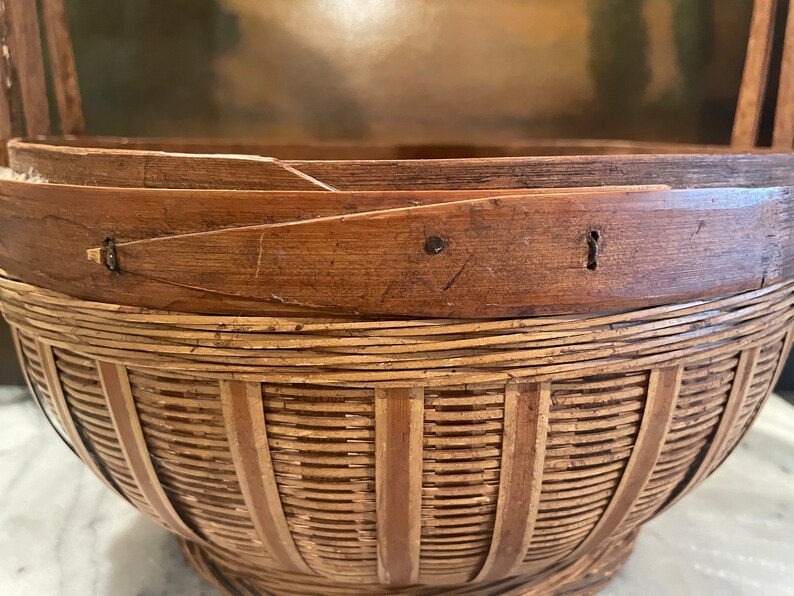 Puede incluir: Una cesta tejida de bamb&uacute; marr&oacute;n y natural con fondo redondeado y una abertura ancha. La cesta tiene un asa a cada lado y est&aacute; hecha de materiales naturales.