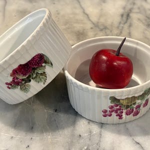 May include: Two white ceramic ramekins with fluted sides, each decorated with a colorful botanical print of raspberries and green leaves. One ramekin is tilted, while the other sits upright containing a small, shiny red apple.