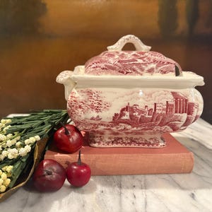 May include: A rectangular ceramic tureen with a lid, featuring a red and white transferware pattern depicting a castle landscape. The piece has ornate handles and a matching lid with a handle, showcasing detailed architectural scenery.