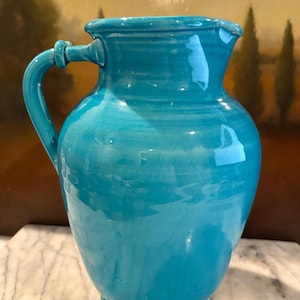 May include: A turquoise ceramic pitcher with a rounded body and a single handle. The pitcher has a smooth, glossy finish.