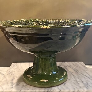 May include: A dark green ceramic pedestal bowl with a glossy finish and a wide, flared rim. The rim features a textured, leaf-like pattern with visible white and light brown distressed edges. The bowl stands on a sturdy, circular base.