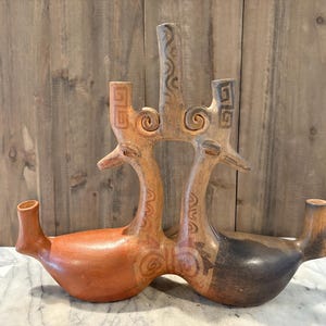 May include: A ceramic vessel with a unique design. The vessel features two bird-like figures joined at the neck, with a central vertical element. The colors include shades of brown, orange, and black. The vessel has a smooth, glossy finish.