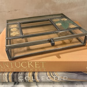 May include: A rectangular glass and metal jewelry box with dried flowers inside, resting on top of two books. The top book is titled "NANTUCKET" in gold lettering. The bottom book is titled "COLLECTED".