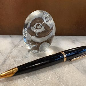 Puede incluir: Un bol&iacute;grafo negro con detalles en color dorado y una punta dorada, colocado frente a una figura de b&uacute;ho de cristal macizo transparente. El bol&iacute;grafo presenta una banda dorada alrededor del centro y un clip dorado en la tapa.